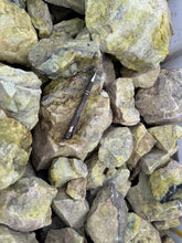 A collection of Serpentine rocks with varying shades of green and a few pieces displaying yellowish hues. The rocks have a rough, textured surface.